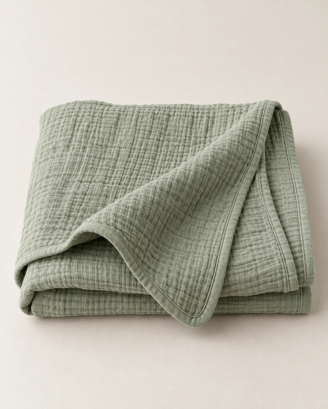 Folded sage cotton muslin wrap with a soft draped corner on a pale tabletop.