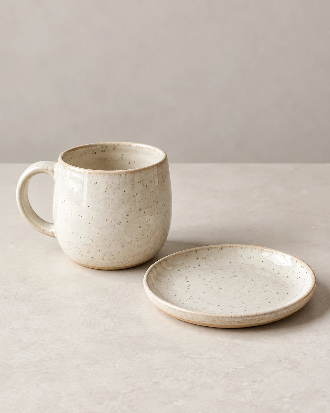 Speckled off-white ceramic mug beside a matching side plate on a softly lit pale surface.